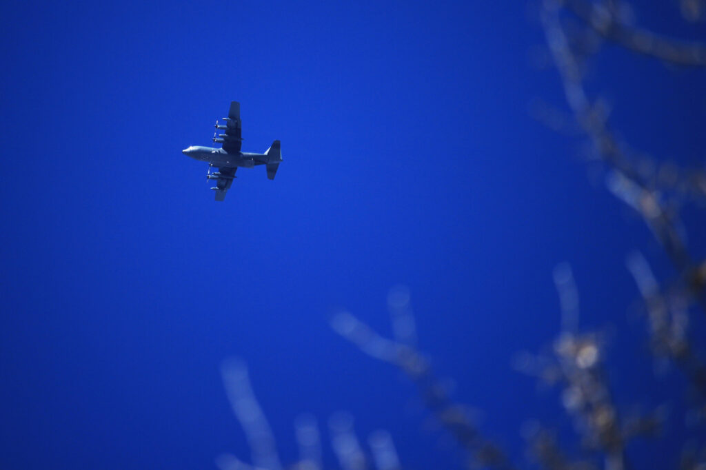 A C-130 Hercules high in the sky, with some bare tree branches in the foreground