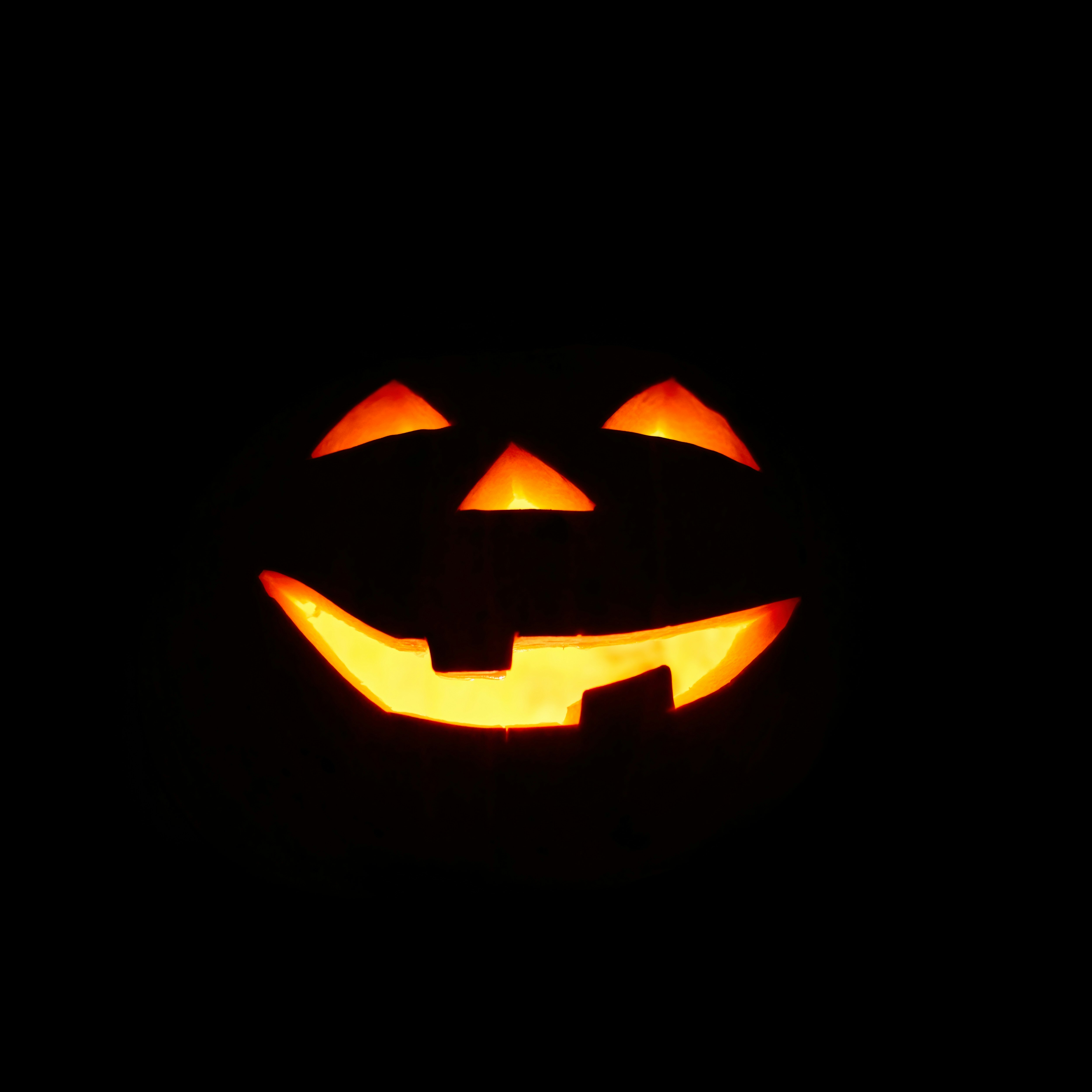 A very dark photo of a Jack-o'-lantern; all we can really see is a glowing set of eyes, nose, and a snaggletoothed smile