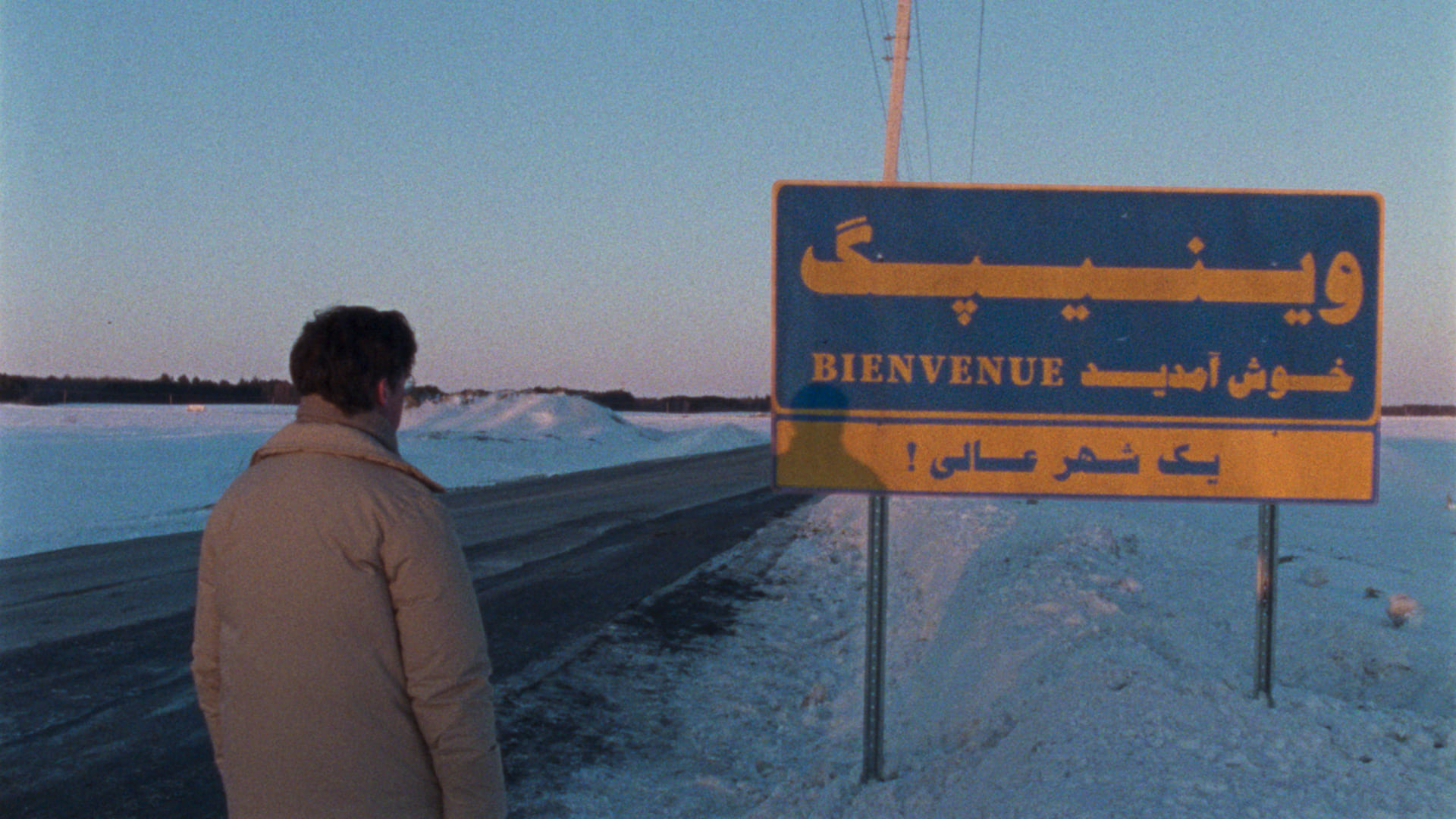 Still from Universal Language: A man stands outside in the winter, looking at a sign that says (in Farsi and French) WELCOME TO WINNIPEG / ONE GREAT CITY